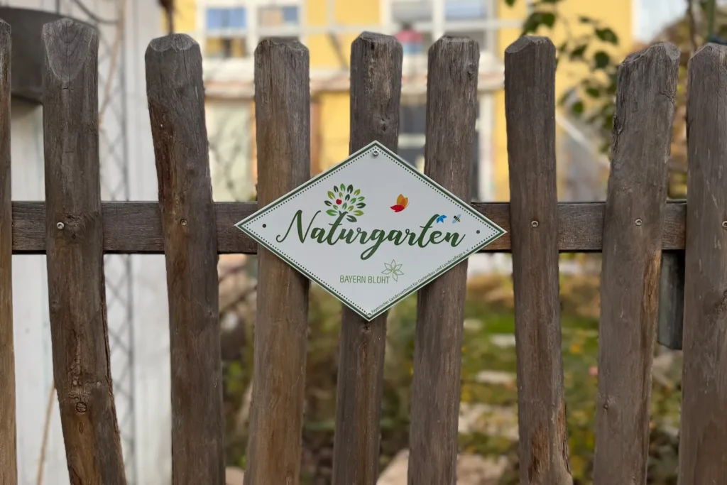 Holzzaun mit einem rautenförmigen Metallschild, auf dem ‚Naturgarten – Bayern blüht‘ steht. Das Schild zeigt ein stilisiertes grünes Bäumchen sowie kleine bunte Schmetterlinge. Im Hintergrund ist ein unscharfer Gartenbereich zu erkennen.