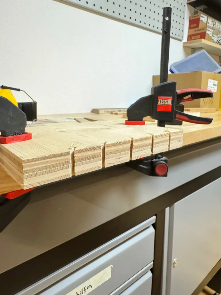 Eine Holzplatte mit mehreren gesägten Nuten. Die Platte wird mit Schraubzwingen gehalten.
