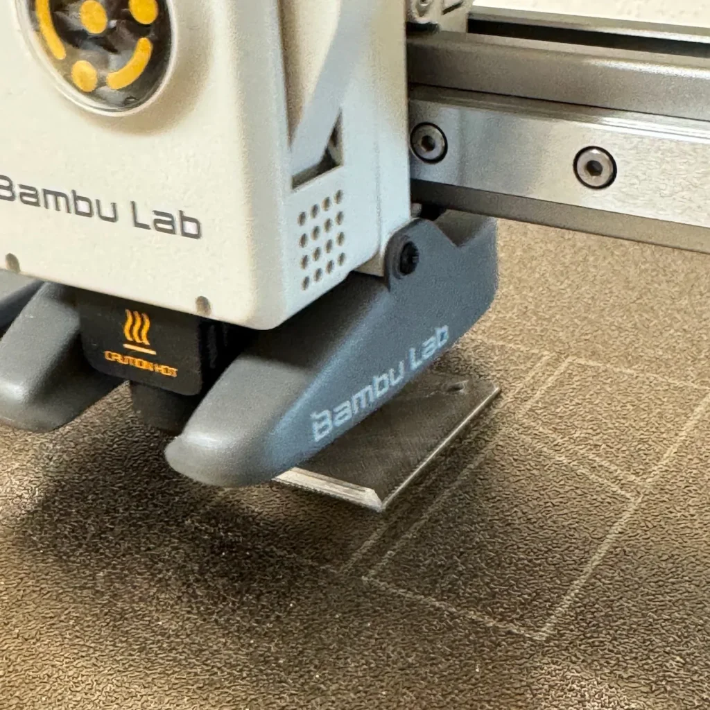Nahaufnahme eines Bambu Lab A1 Mini 3D-Druckkopfs beim Drucken einer transparenten Wachsmottenklappe auf einer strukturierten Druckplatte.