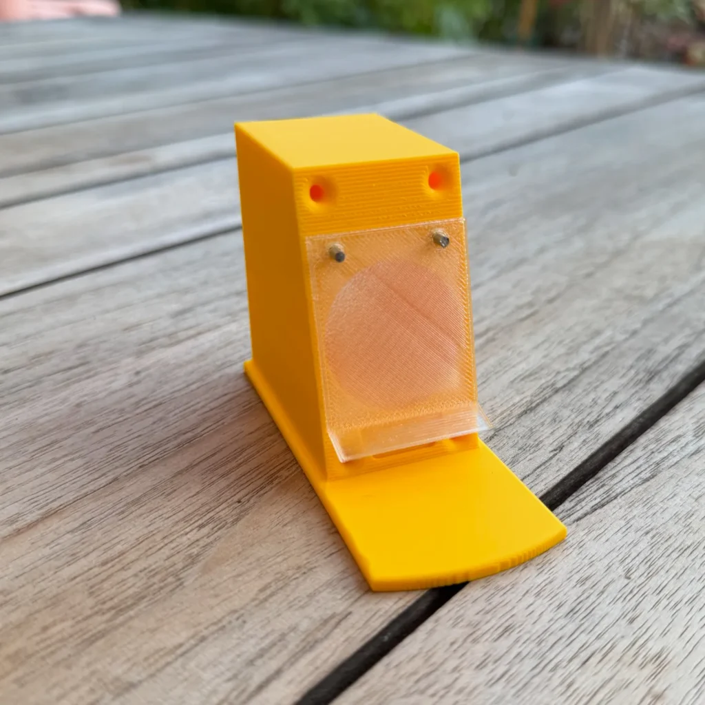 Gelber Vorbau mit montierter transparenter Klappe, befestigt mit zwei kleinen Nägeln.