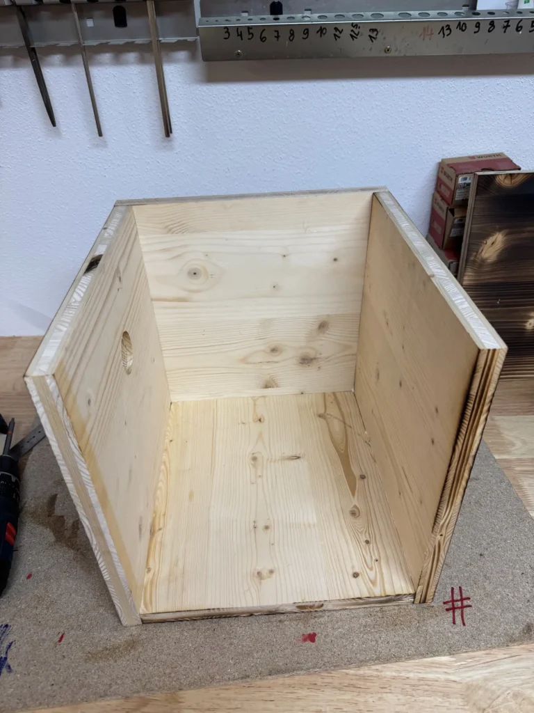 Teilweise zusammengebauter Holzkasten aus hellen Platten.