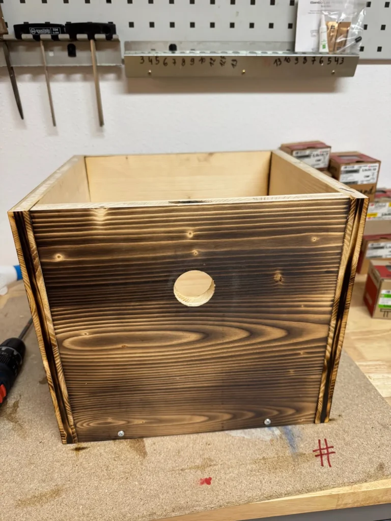 Fertig montierter Holzkasten mit geflammter Oberfläche und runder Öffnung auf einer Werkbank.