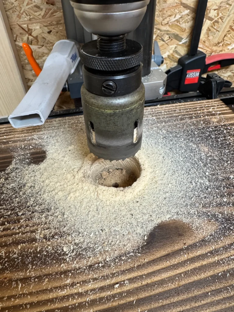 Bohrmaschine mit Lochsäge bohrt ein rundes Loch in eine geflammte Holzplatte, Holzspäne ringsum.