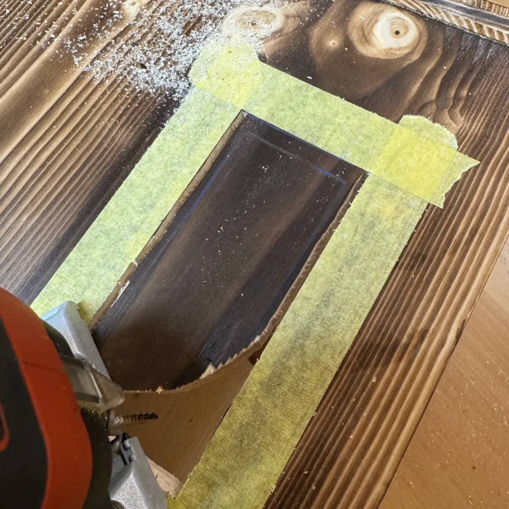 Ausschnitt in einer geflammten Holzplatte, mit gelbem Klebeband markiert und mit einer Stichsäge ausgeschnitten.