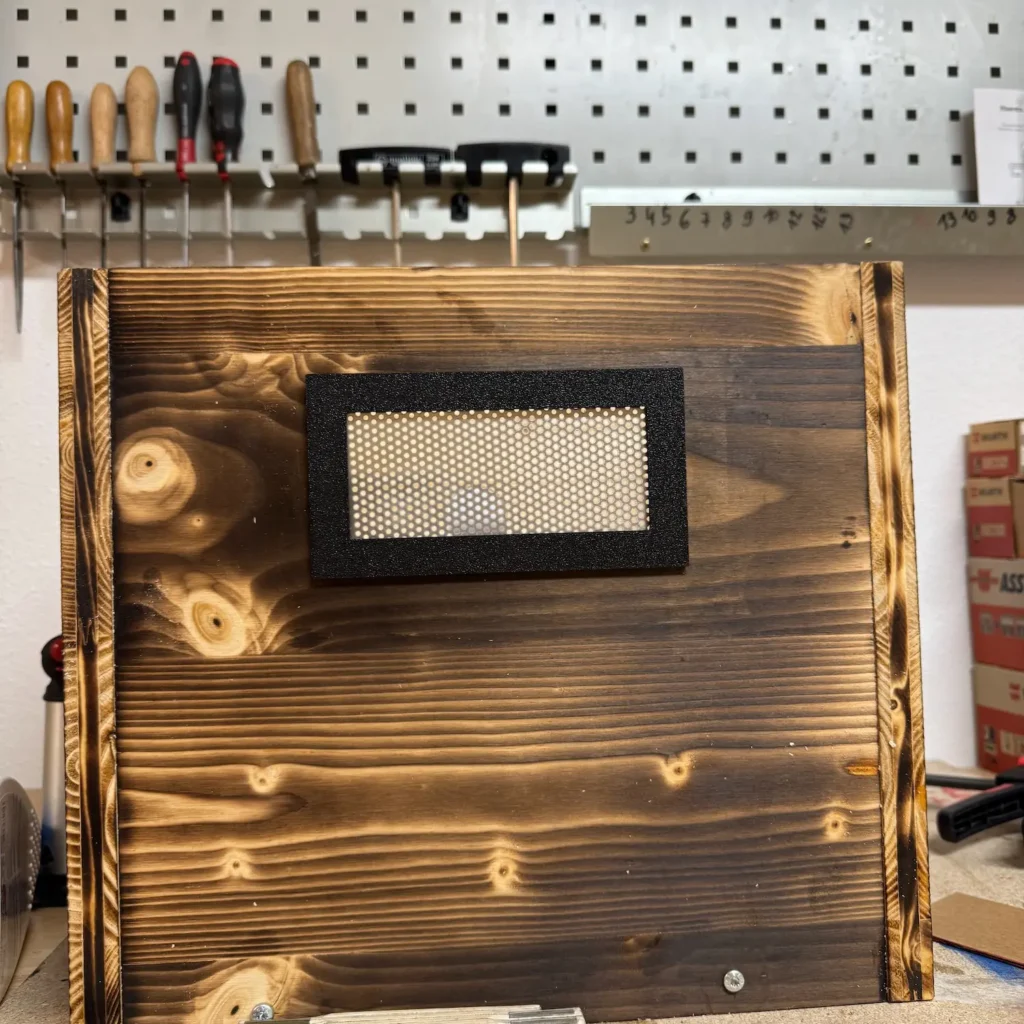 Schwarzer Lüftungsrahmen mit Metallgitter auf einer geflammten Holzplatte befestigt.