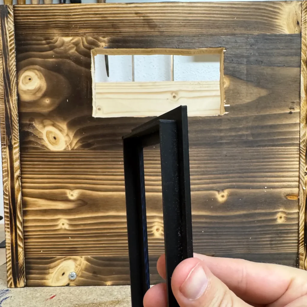 Hand hält einen schwarzen 3D-gedruckten Rahmen vor eine rechteckige Aussparung in einer geflammten Holzplatte.