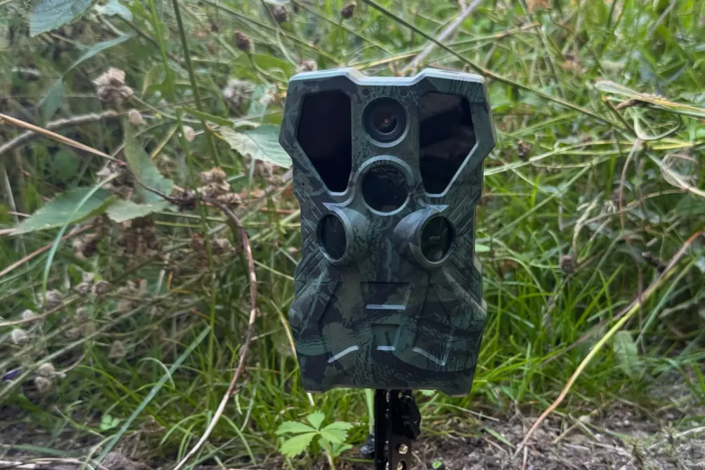 Wildkamera PetTec Pet Cam Trail im Tarngehäuse, auf einem Stativ im hohen Gras aufgestellt.