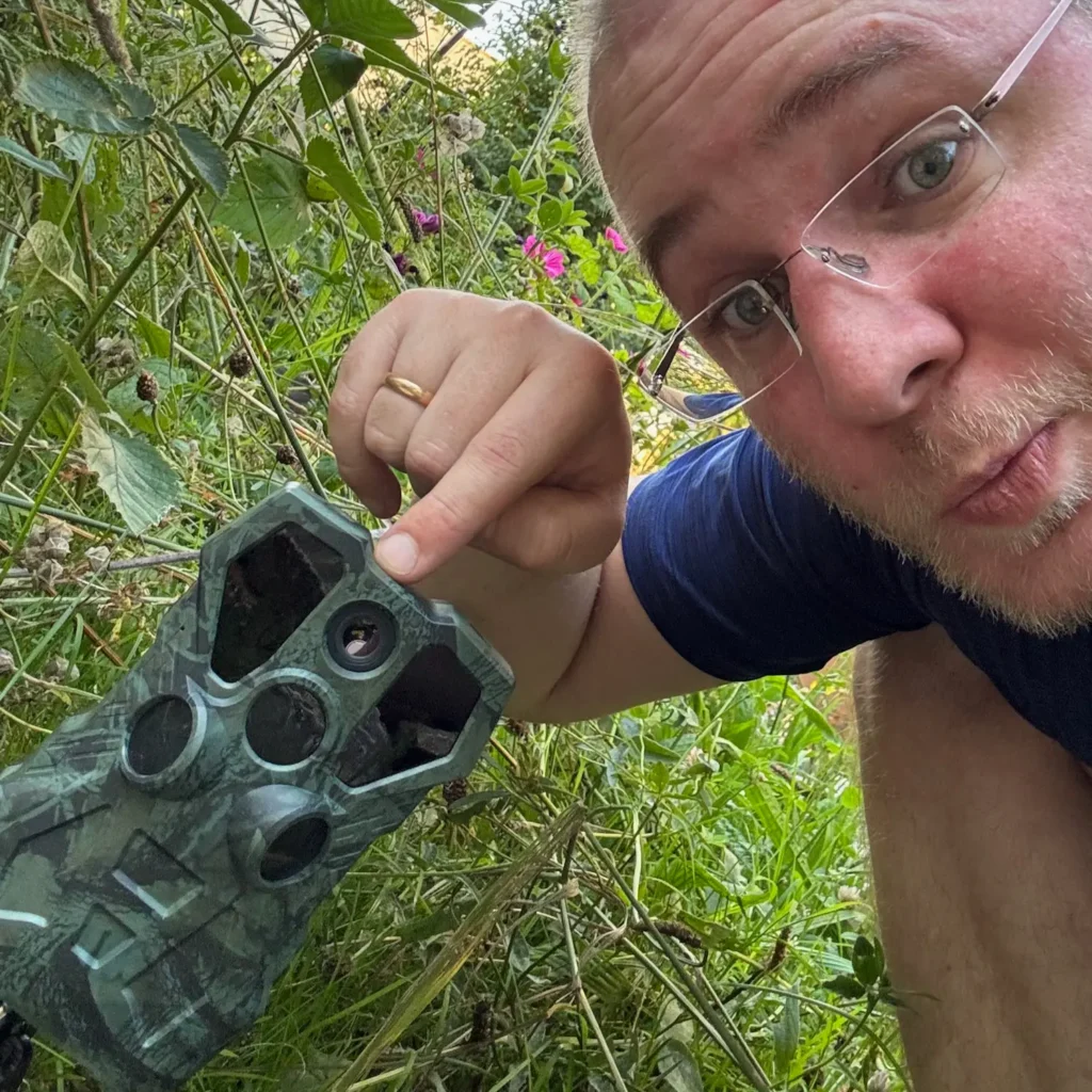 Selfie eines Mannes der im Garten kniet und mit dem Finger auf einer Wildtierkamera zeigt.
