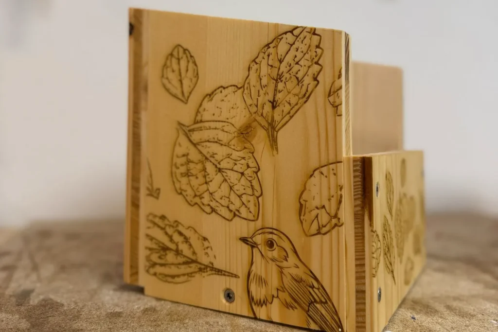 Auf der Seitenwand des Halbhöhlen-Nistkastens ist ein kleiner Vogel zusammen mit Blättern graviert.