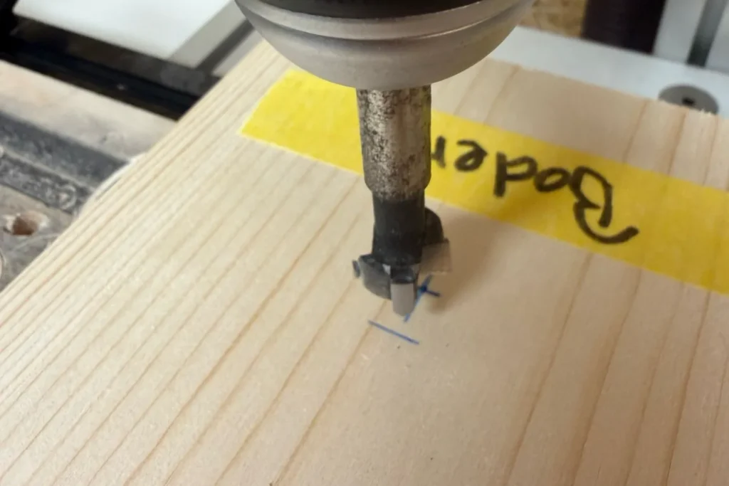 Mit einem Forstnerbohrer wird ein Wasserablauf in die Bodenplatte des Halbhöhlen-Nistkastens gebohrt.