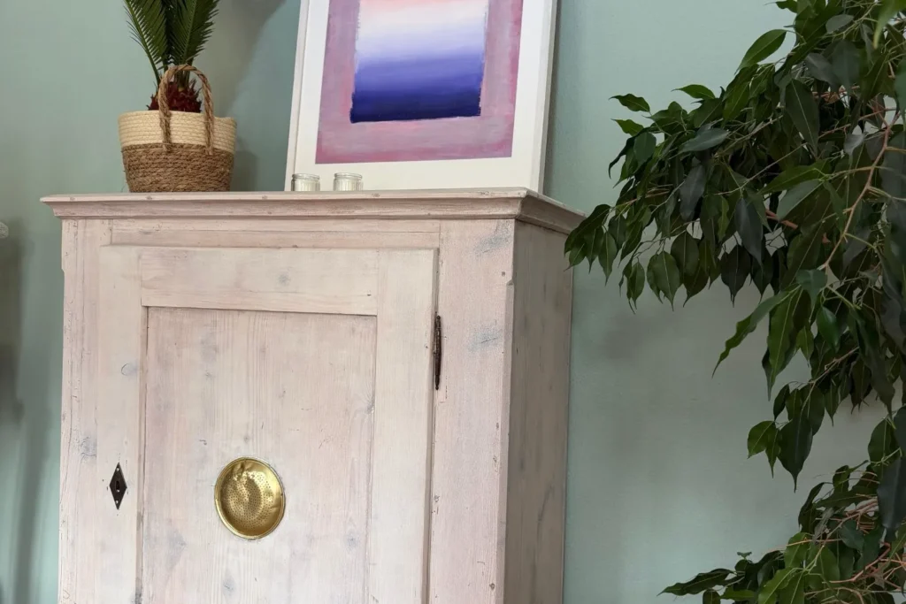 Restaurierter Brotschrank mit kalkweißer Lasur und goldenem Lüftungsgitter, dekoriert mit Pflanze, Bild und Kerzen im Wohnzimmer.