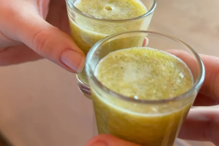 Zwei Hände stoßen mit selbstgemachten grünen Ingwer-Shots in kleinen Gläsern an.