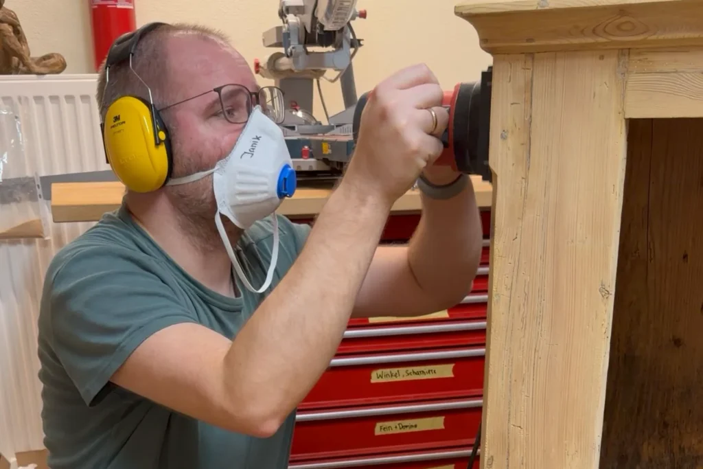 Person beim Schleifen eines alten Holzschranks, mit Atemschutz und Gehörschutz