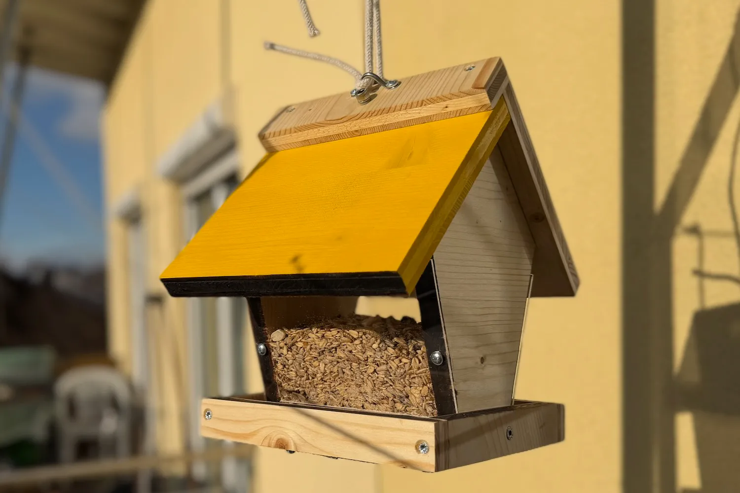 Selbstgebautes Vogelfutterhaus aus Holz mit gelb gestrichenem Dach und Lochblechboden, aufgehängt im Garten