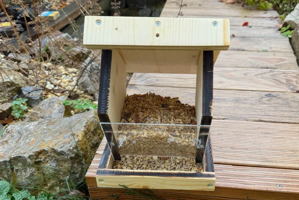 Frontansicht eines selbstgebauten Vogelfutterhauses mit Plexiglasscheibe und eingefülltem Körnerfutter.