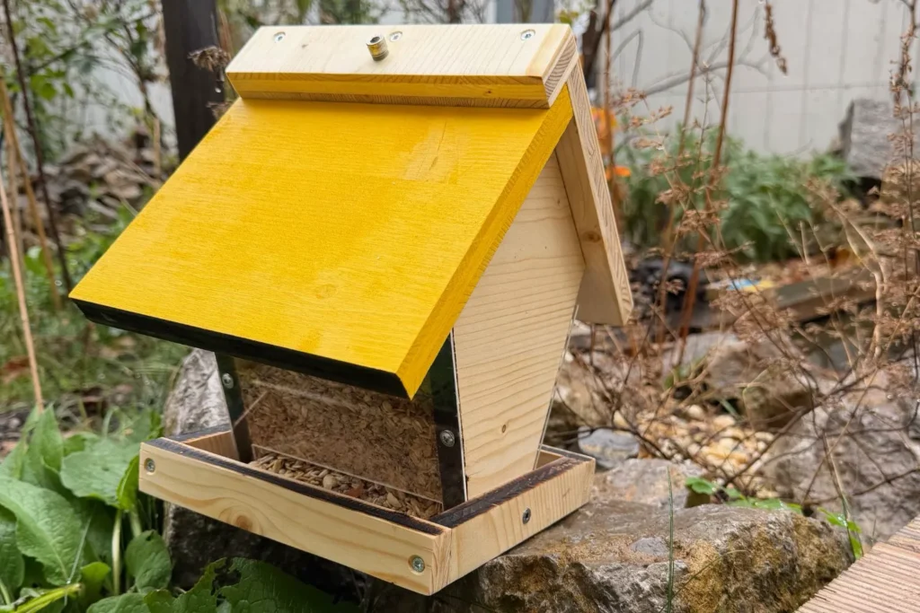 Selbstgebautes Vogelfutterhaus mit gelbem Dach steht im Garten auf einem Stein, umgeben von Pflanzen.