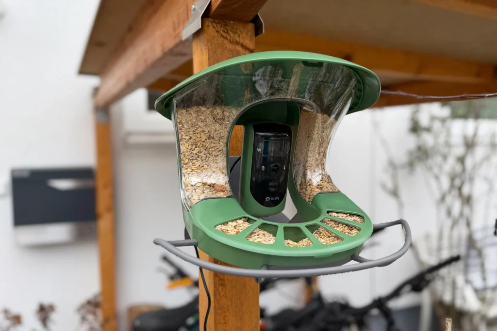 Birdfy Vogelfutterstation mit integrierter Kamera, unter einem Holzvordach montiert und mit Körnerfutter befüllt.