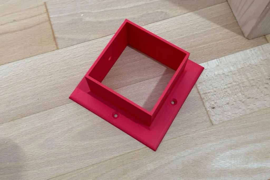 Roter Pfostenschuh aus dem 3D-Druck zur Befestigung eines Holzpfostens auf dem Boden.