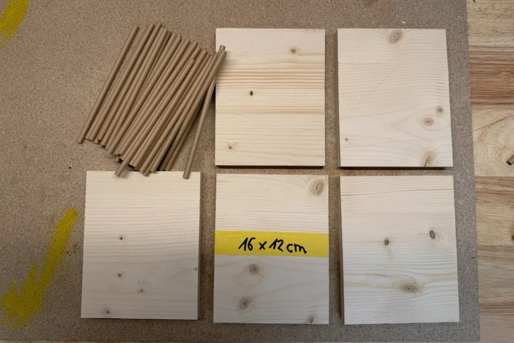 Materialübersicht für eine selbstgebaute Wildbienen-Nisthilfe aus Fichtenholz mit zugeschnittenen Holzplatten und Nisthülsen.